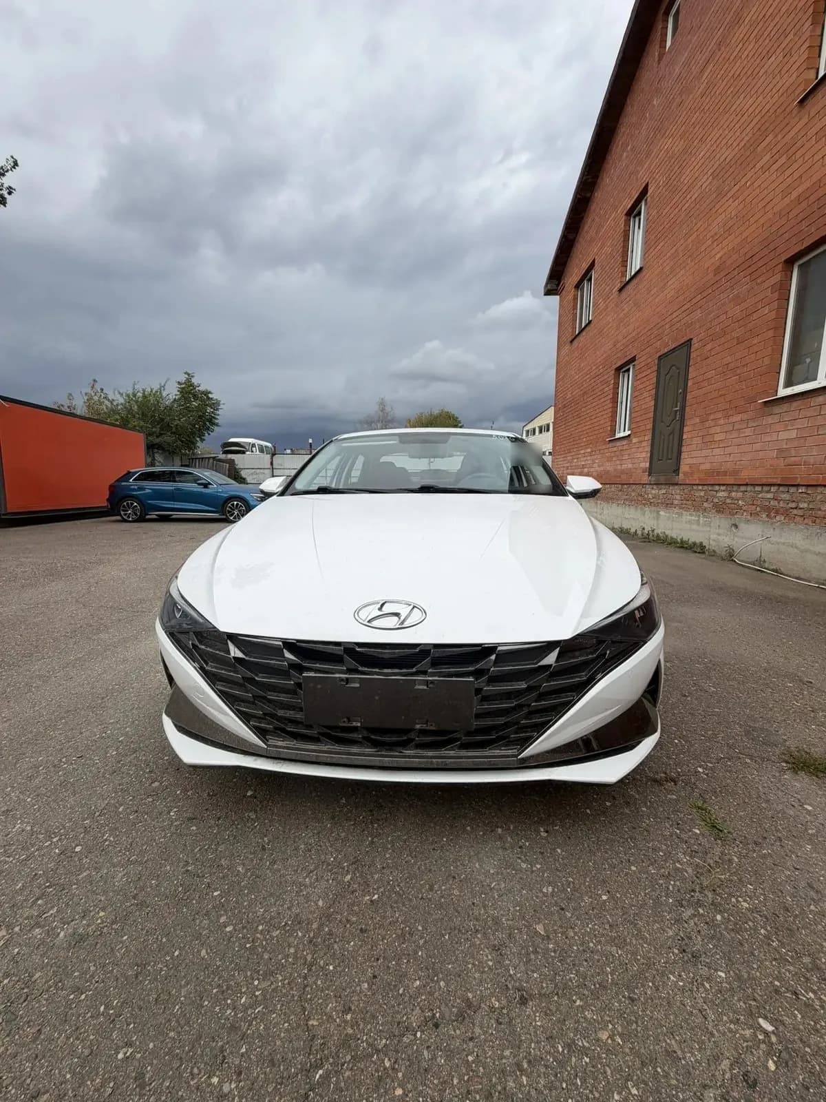 Белая Hyundai Elantra спереди, общий план площадки: слева вагончик и синяя машина, справа кирпичное здание