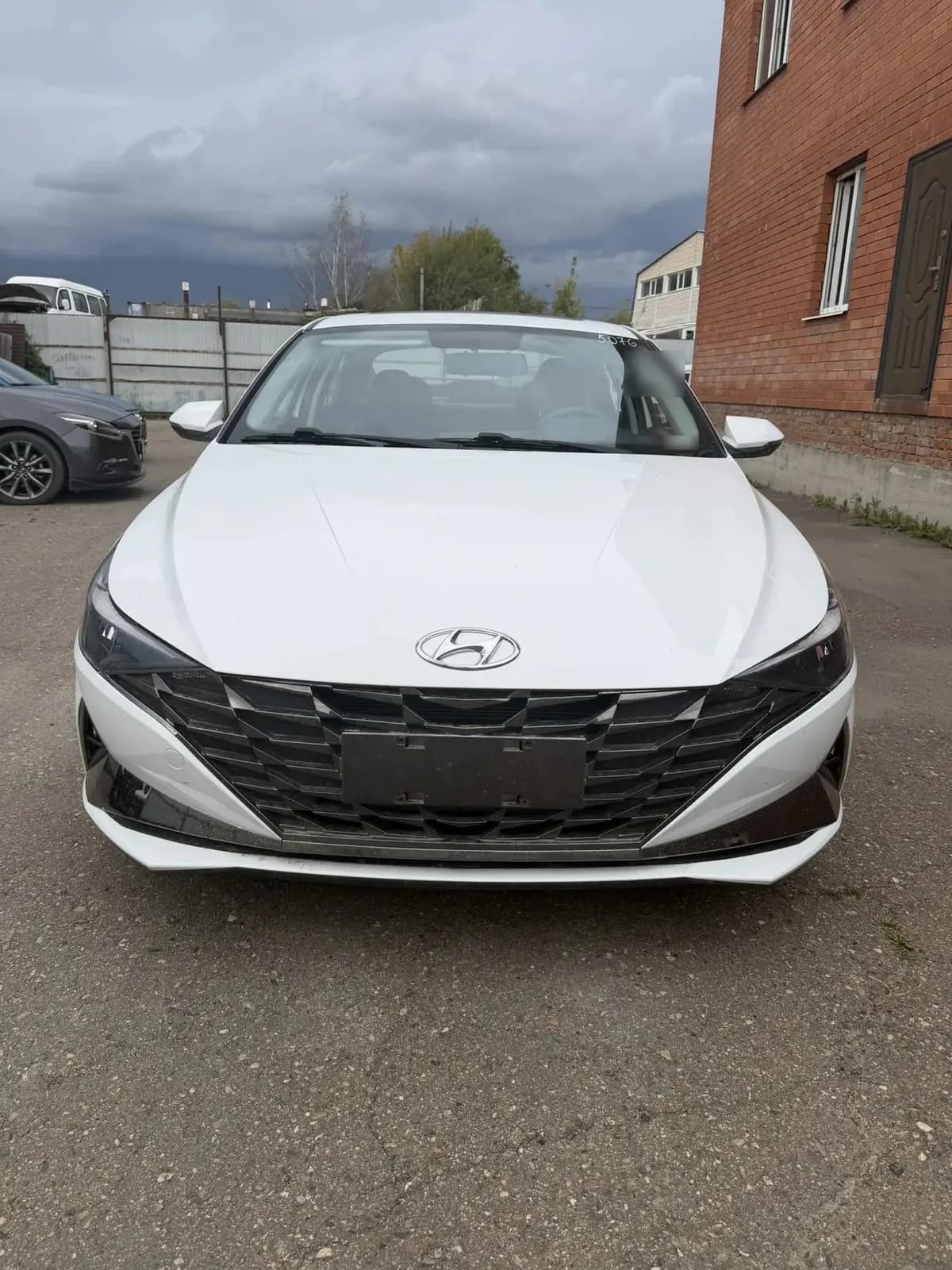 Белая Hyundai Elantra спереди на площадке: слева стоят другие машины, справа кирпичное здание