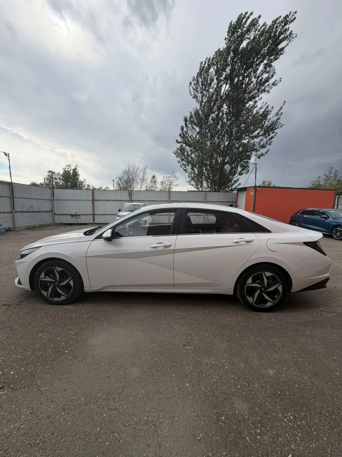 Белая Hyundai Elantra, левый борт: на заднем плане оранжевый вагончик и другие машины