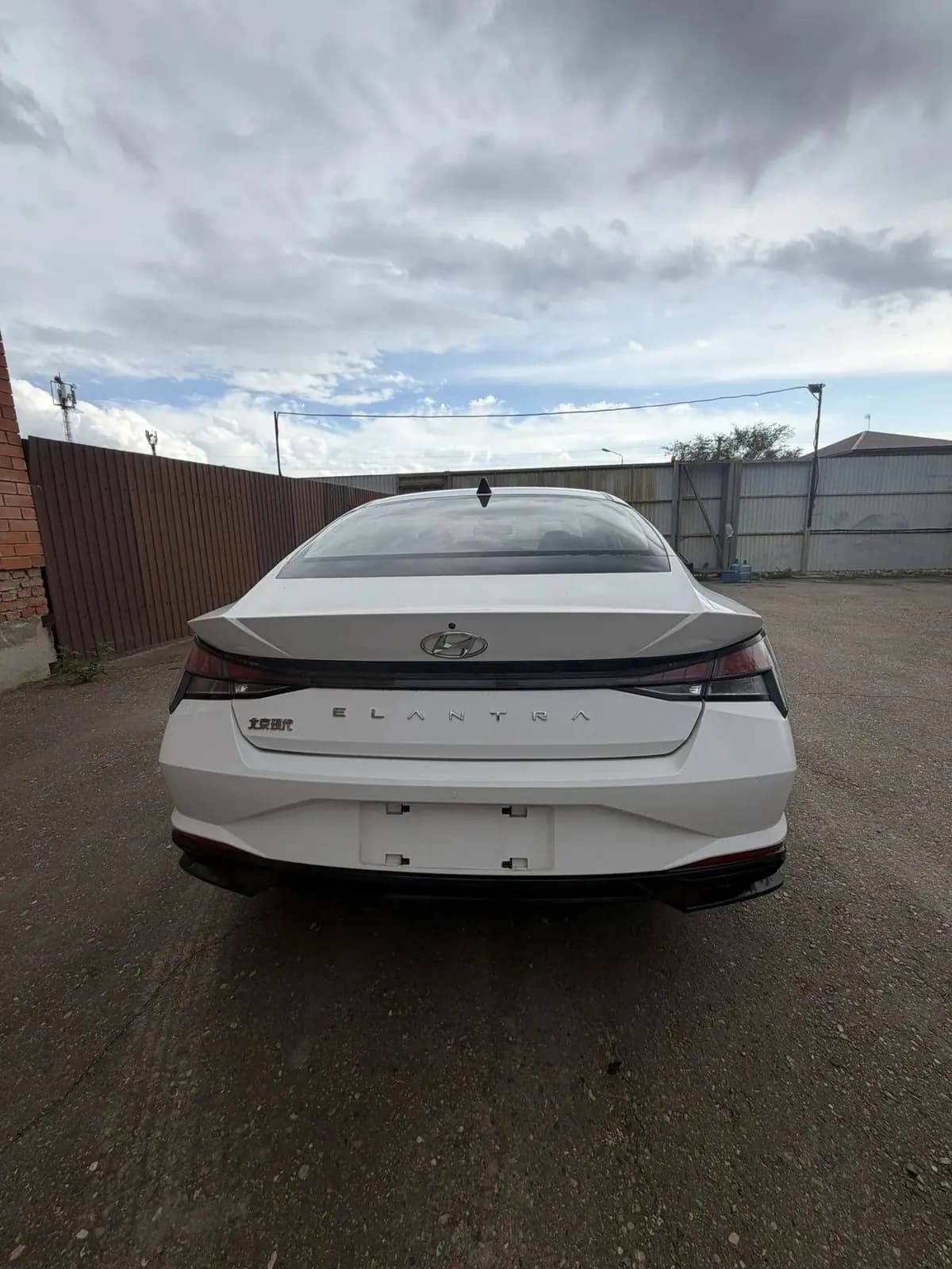 Белая Hyundai Elantra сзади, общий план площадки у коричневого забора