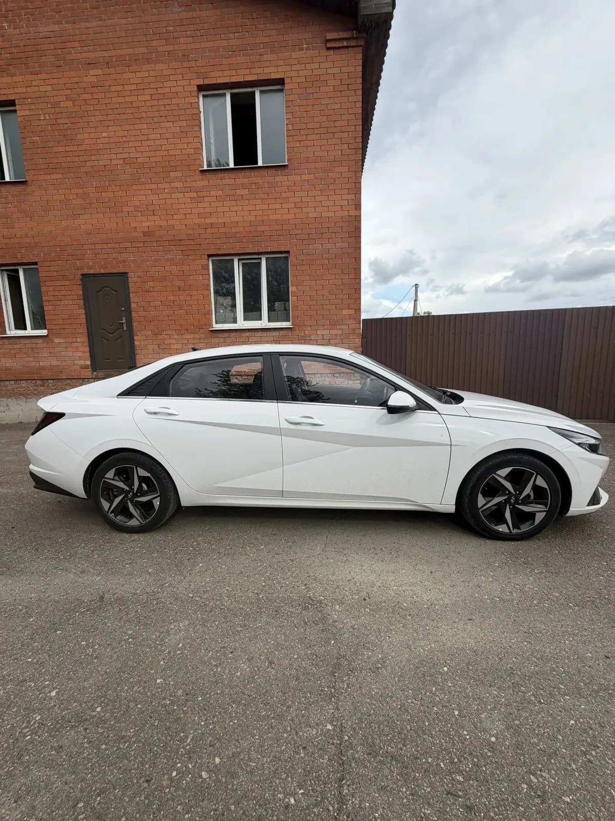 Белая Hyundai Elantra, правый борт: рядом кирпичное здание и коричневый забор